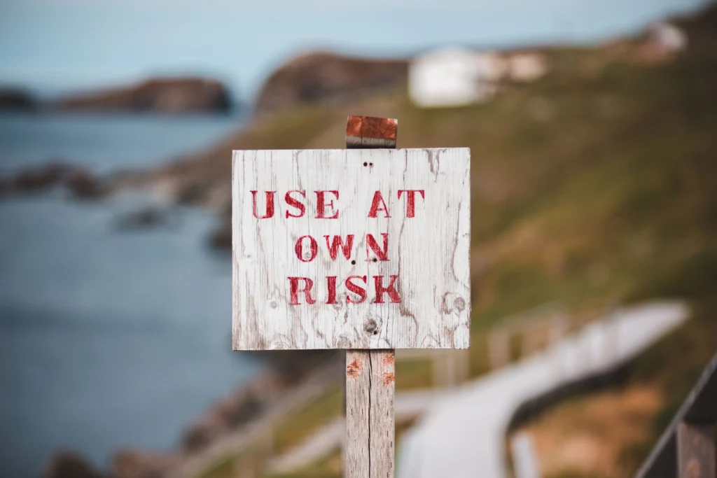 Close up of a wooden sign with red text stating USE AT OWN RISK with a blurred ocean coastline background